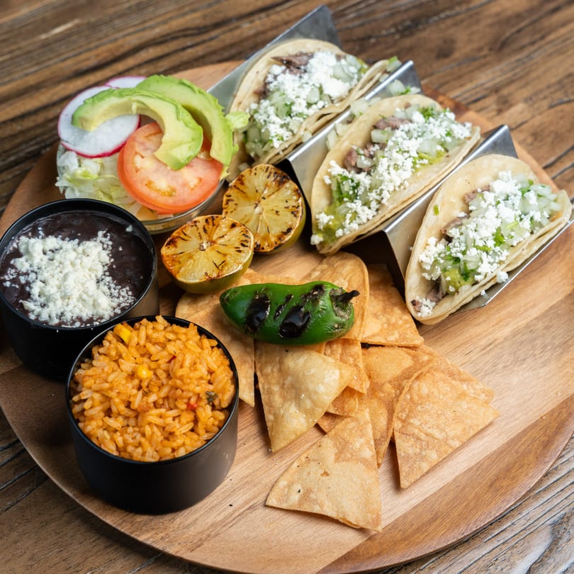 Mexican Mariachi Grill's tacos with sides of rice, beans, salad, chips, and grilled lime on a wooden platter.