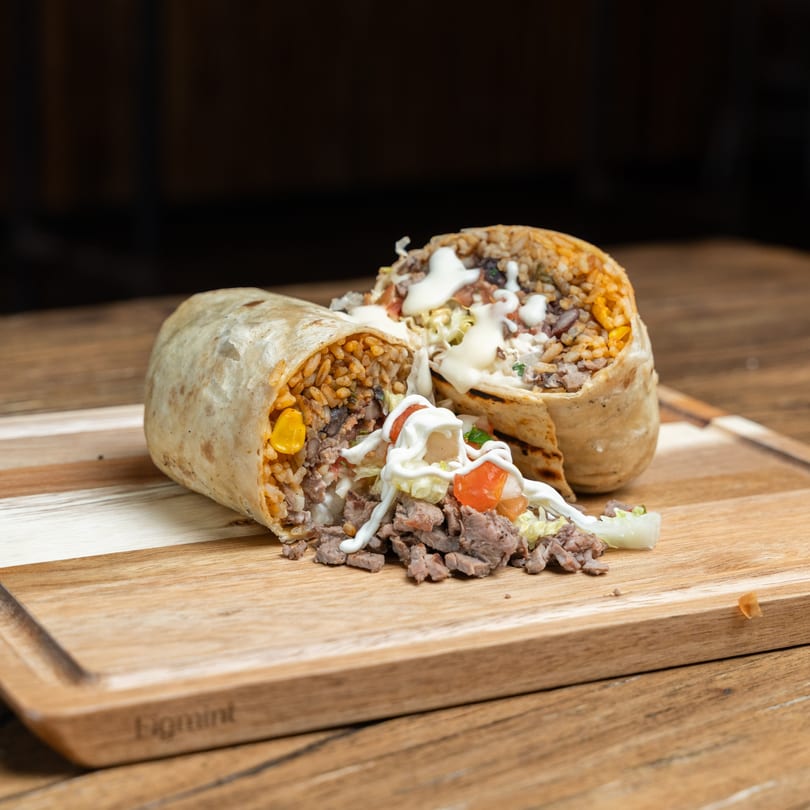 Two Mexican burritos, one cut open, filled with meat, rice, corn, and drizzled with white sauce.