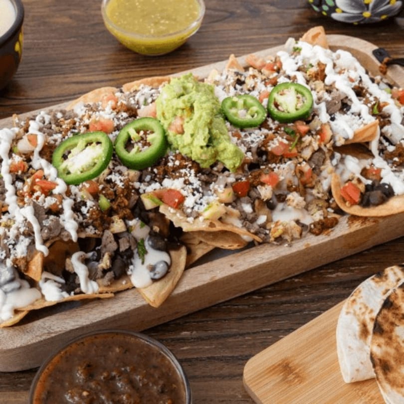 Loaded Mexican nachos with meat, beans, cheese, guacamole, sour cream, and jalapeños on a wooden board.