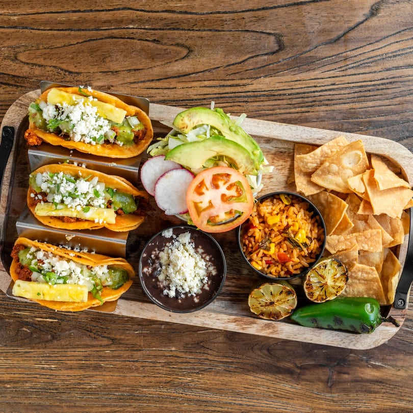 Three Mexican tacos with pineapple, cheese, and salsa, served with rice, beans, chips, and grilled lime.