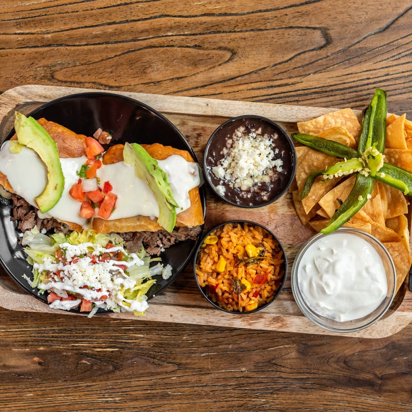Mexican Mariachi Grill's carne asada with rice, beans, chips, and sour cream on a wooden tray.
